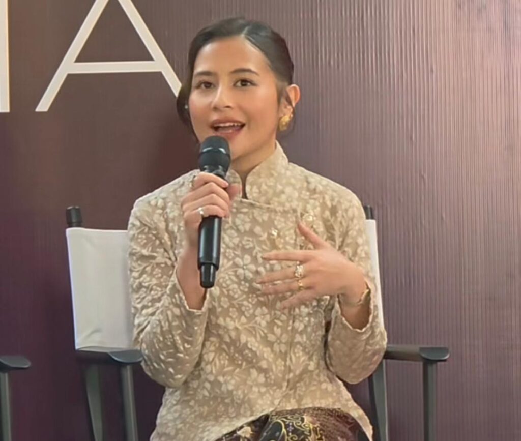 Whatsapp image 20260619 at 14 14 08 catrawarta A woman in a beige floral traditional outfit speaks into a handheld microphone on stage gesturing with her other hand